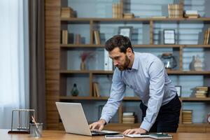 Focused mature businessman reviewing documents at his laptop in a well-organized, modern home office setting. photo