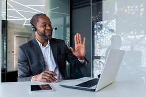 Mature experienced man with headset phone inside office at workplace, smiling talking remotely and advising customers, african american customer support worker, using call and laptop. photo