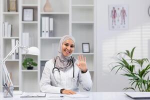 Smiling female doctor in hijab waves at the camera during a call in a well-lit, modern office setting, displaying warmth and professionalism. photo