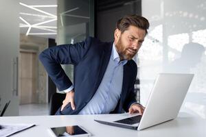 Young male businessman sitting in the office at the desk and holding his back, suffering from pain and spasms. photo