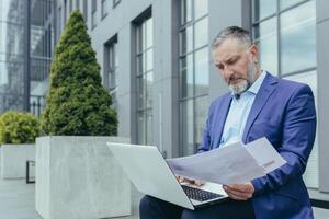 grave mayor hermoso exitoso masculino empresario trabajos con ordenador portátil y documentos, concluye en línea contrato con clientela. sentado en un banco cerca el oficina centro. foto