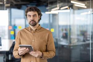 Focused businessman with digital tablet standing in contemporary office space, looking thoughtfully into distance. photo