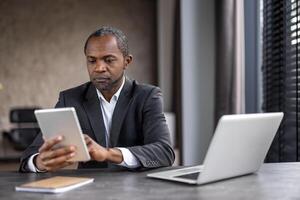 A man in a suit is sitting at a desk with a tablet and a laptop. He is focused on the tablet, possibly checking emails or browsing the internet. Concept of productivity and concentration photo