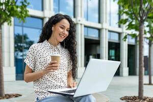 un alegre joven mujer se sienta fuera de con un ordenador portátil y café, tomando un agradable descanso desde trabajo mientras vistiendo auriculares. foto