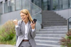 Successful mature joyful businesswoman received online notification message with good results achievement on phone, woman boss uses app on smartphone, walks outside office building in business suit. photo