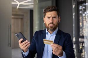 Close-up portrait of serious and confused young businessman having problems with credit card and online application on phone, looking worriedly at camera. photo