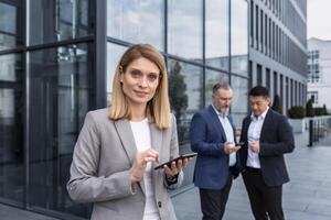 retrato de exitoso mayor mujer empresario y especialista, programa gerente con tableta computadora fuera de oficina edificio sonriente y mirando a cámara, profesional programador equipo líder. foto