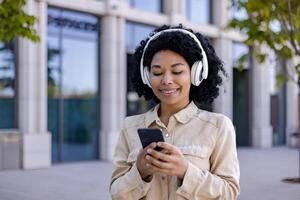 joven hermosa africano americano mujer caminando en el ciudad vistiendo auriculares sonriente escuchando a música y audio libros, mujer utilizando teléfono inteligente solicitud para en línea podcasts foto