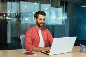 Successful businessman in red shirt happily working with laptop inside office, mature man with beard at workplace typing on keyboard smiling satisfied with work results and achievement. photo