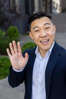 Vertical close-up photo of a young Asian businessman talking on a call, greeting and waving at the camera.