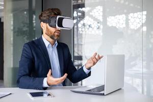 Businessman in suit using VR headset at desk in modern office, gesturing with hands, exploring virtual reality. photo