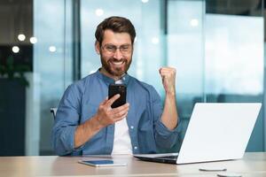 contento joven hermoso hombre persona de libre dedicación jugando en línea juegos en el teléfono a trabajar. levanta su mano, muestra Sí, ganado. sentado en el oficina a el escritorio. foto