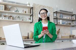 hermosa joven mujer en lentes y verde camisa relajante a hogar en cocina, escuchando música foto