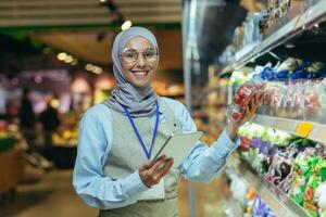 Portrait of happy and smiling seller woman in hijab, muslim woman with tablet smiling and looking at camera, saleswoman near field with vegetables and salad products chooses and sifts term photo