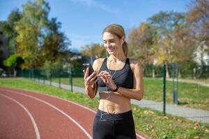 A smiling woman in athletic wear prepares for a run at the track by choosing music or an app on her smartphone. photo