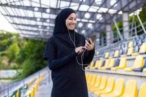 Happy woman in hijab and headphones doing sports in football arena, holding phone and listening to music. photo
