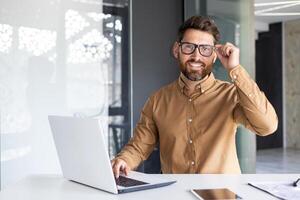 retrato de exitoso hombre a lugar de trabajo dentro oficina, experimentado sonriente empresario en camisa sonriente y mirando a cámara, programador desarrollando nuevo software con ordenador portátil. foto