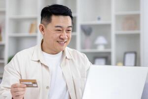 Joyful successful asian businessman working from bright home office, man using bank credit card and laptop for online shopping in online store, and booking services remotely. photo