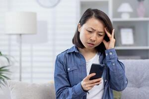 joven asiático mujer trastornado sentado a hogar, mujer tiene malo Noticias en línea en teléfono sentado en sofá en vivo habitación, trastornado y decepcionado. foto