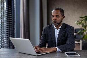 Focused male executive in formal attire typing on a laptop at a modern workspace with natural light and tablet on desk. photo