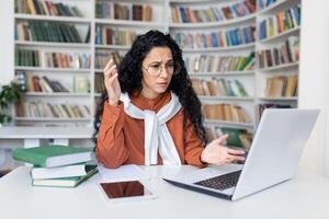 Front view of confused female with caucasian appearance working remotely from home office with designer shelves with books on background. Woman feeling perplexed while using modern laptop indoors. photo