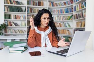 frente ver de confuso hembra con caucásico apariencia trabajando remotamente desde hogar oficina con diseñador estantería con libros en antecedentes. mujer sensación perplejo mientras utilizando moderno ordenador portátil adentro. foto