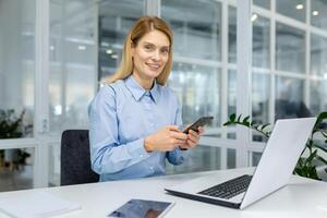 Modern professional woman using a smartphone at her workspace with a laptop, tablet, and documents on a white desk. photo