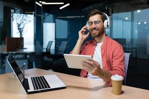 A young freelancer, female student, programmer sits in the office at the table with a laptop, listens to music in headphones, holds tablet in hands. He closed his eyes, rests, relaxes, dances, sings. photo