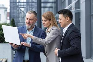 Successful business team, three colleagues businessman and businesswoman outside office building discussing current plans and management, looking at laptop screen, discussing and consulting photo