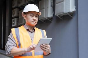 joven asiático masculino ingeniero, construcción trabajador en pie fuera de edificio y utilizando tableta. foto