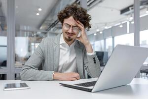 Exhausted young businessman at his desk in a bright office rubbing his temples, feeling stressed while working on a laptop. photo