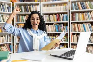 alegre mujer en lentes celebra como ella recibe un positivo postal mensaje, levantamiento su puño en triunfo a un biblioteca. foto