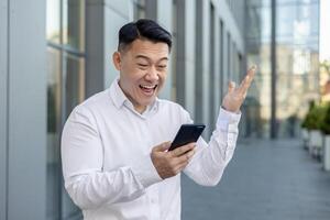 A professional businessman in a white shirt uses a smartphone while making a hand gesture against an urban backdrop. photo