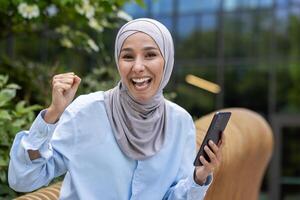 joven hermosa musulmán mujer en hijab sentado en banco fuera de oficina edificio, mujer de negocios mirando a cámara, recibido en línea mensaje, victoria éxito, participación mano arriba celebrando triunfo foto