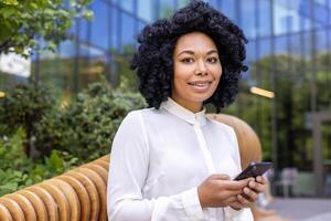 retrato de un sonriente joven africano americano mujer de negocios sentado en un banco fuera de un oficina edificio, participación un móvil teléfono y mirando a el cámara. de cerca foto. foto