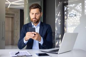 concentrado y grave joven empresario trabajando en el oficina en un computadora portátil, sentado a el mesa y mecanografía un mensaje, mensajes de texto en un móvil teléfono. foto