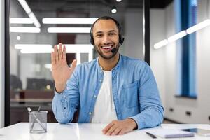 sonriente joven africano americano hombre en auriculares sentado en oficina en frente de cámara, hablando y ondulación mano a cámara. foto