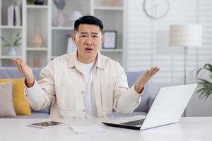 Worried Asian man working at home on laptop, looking confused at camera, spreading hands. Financial problems, broken device, remote work. photo