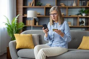 Anxious mature woman sitting on sofa, holding credit card and phone, looking at screen with concern, suspecting a fraudulent transaction. photo