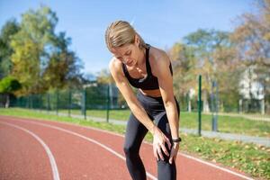 Fit woman in athletic wear rests with hands on knees after running on track field, displaying workout fatigue and determination. photo