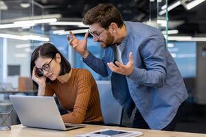 agresivo masculino jefe en mezclilla camisa extensión manos y Gritando a irritado hembra empleado por trabajando escritorio. estresado mujer sentado a oficina en frente de ordenador portátil y tomando crítica desde director. foto