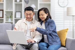 Happy asian family, couple in love man and woman together at home sitting on sofa happy in living room choosing products in online store using laptop spending time together. photo