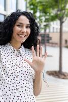 Cheerful woman in polka dot shirt waving hello outdoors, with a bright smile and urban background, conveying friendliness. photo