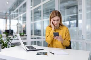 maduro experimentado negocio mujer utilizando aplicación en teléfono en línea sentado dentro oficina a lugar de trabajo con computadora portátil, hembra trabajador conmocionado por malo Noticias leyendo en línea mensaje desde teléfono inteligente foto