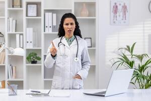 latín americano joven hembra médico en pie en clínica oficina cerca trabajando escritorio, mirando seriamente a cámara y demostración dedo amenazante gesto. foto