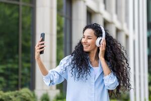 contento joven mujer utilizando teléfono mientras caminando en el ciudad de cerca, Hispano mujer en auriculares utilizando teléfono inteligente aplicación a escucha a música, alegremente canto y baile. foto