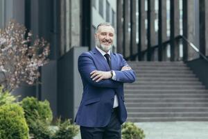 retrato de un exitoso mayor masculino empresario en un negocio traje y Corbata en pie con confianza cerca el oficina centrar en lleno altura, brazos cruzado, sonriente. foto