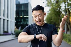 de cerca foto. joven asiático masculino deportista en pie en ciudad calle después corriendo y mirando a inteligente reloj en mano, contento con resultado. foto