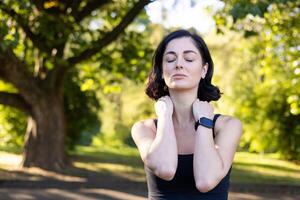 Close-up photo of young tired woman doing sports and exercising outdoors, standing exhausted with closed eyes, holding hands on neck, doing massage and resting.