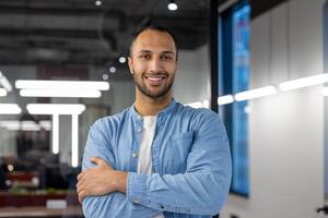 retrato de un joven Hispano masculino programador y desarrollador en pie en el oficina, cruce su brazos terminado su cofre y mirando, a el cámara con un sonrisa. foto